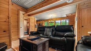 En stuga med träpanel på insidan, med en svart lädersoffa, ett soffbord och ett köksområde som syns genom en öppen dörr.