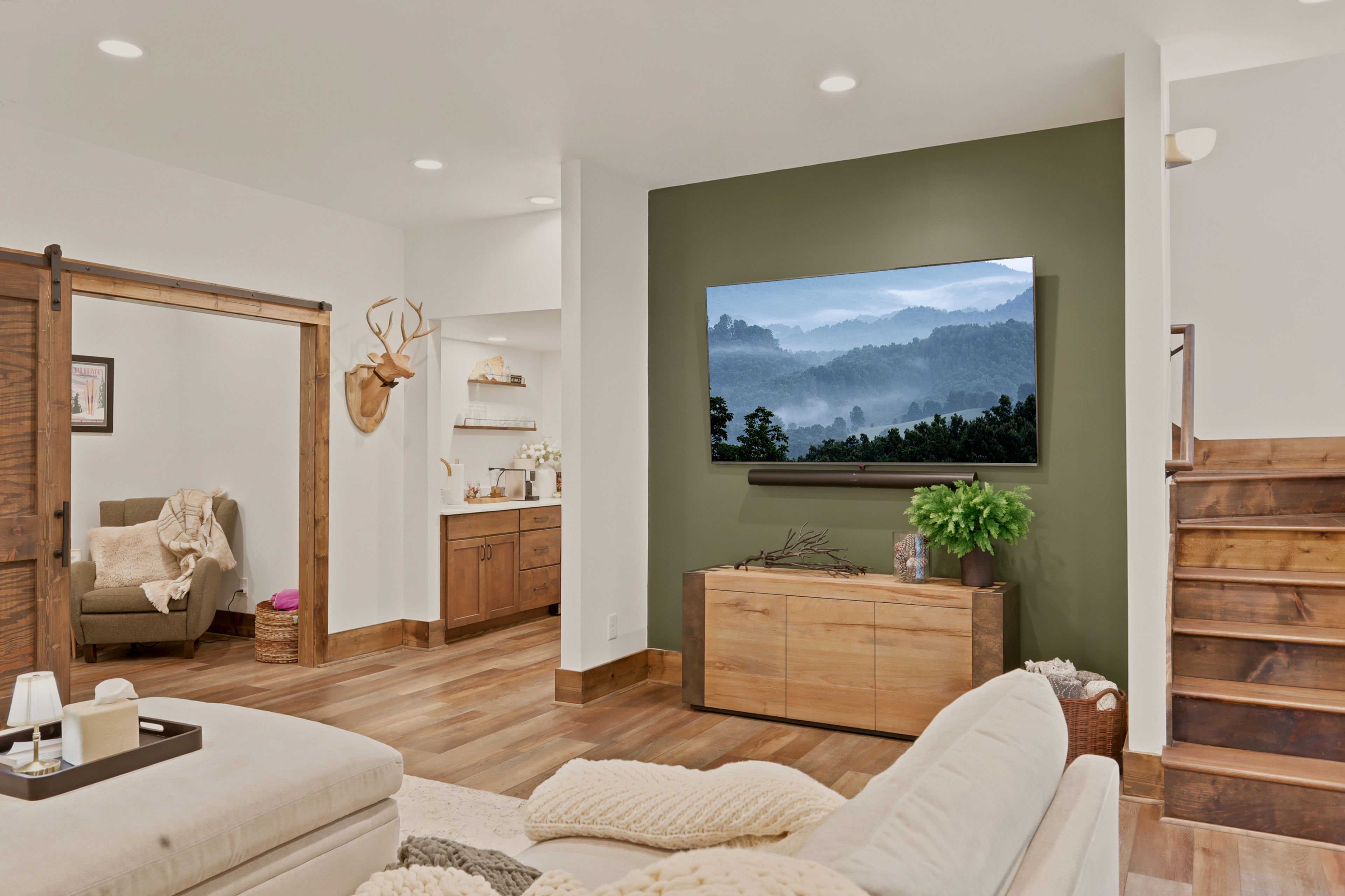 A modern living room with a large flat-screen TV mounted on a green wall, wooden furniture, and a cozy seating area.