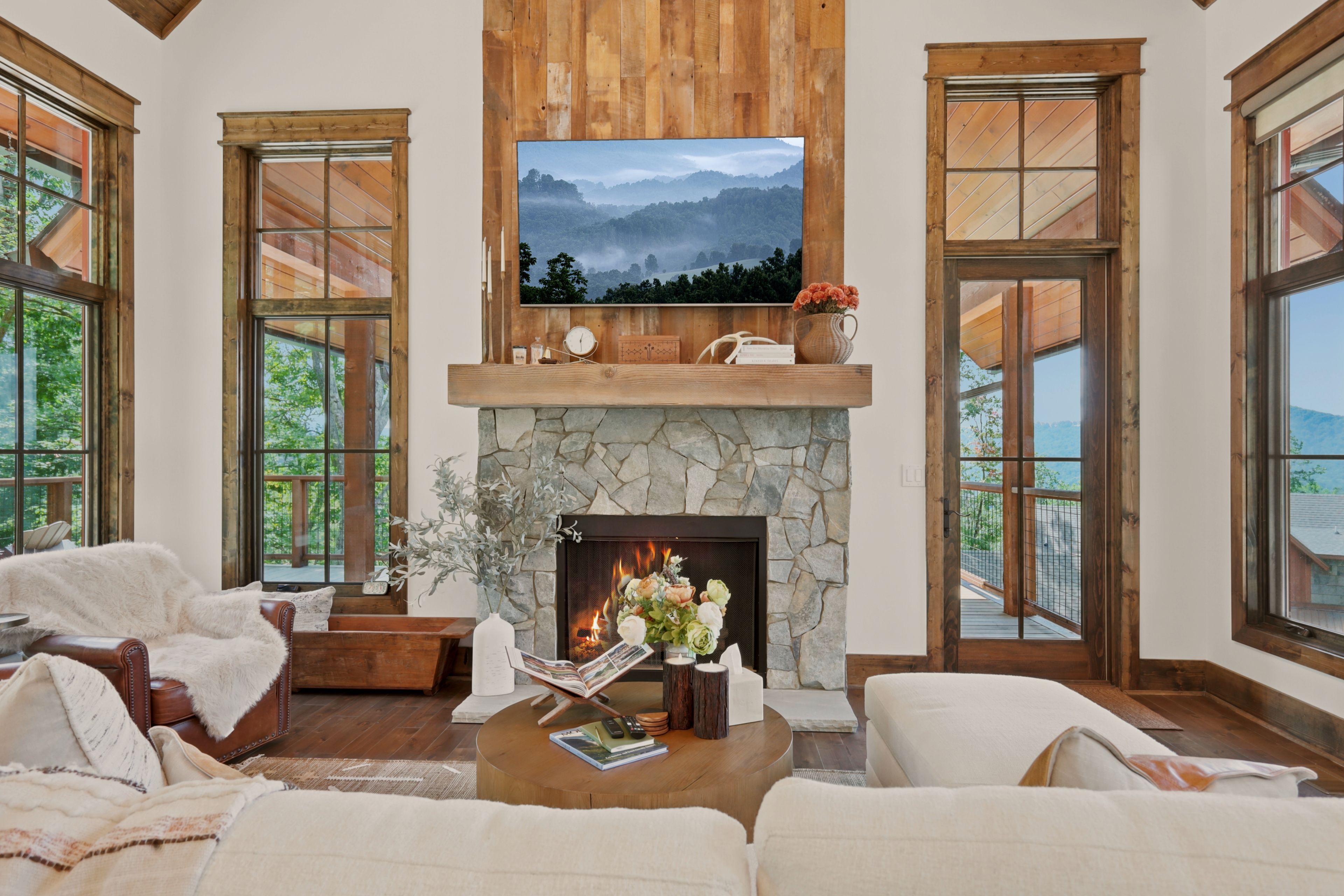 A spacious living room with a stone fireplace, a mounted television, and large windows offering a view of the outdoors.