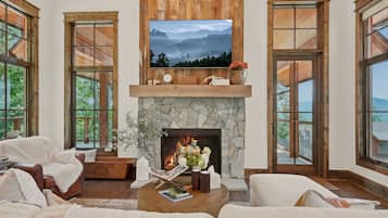 A spacious living room with a stone fireplace, a mounted television, and large windows offering a view of the outdoors.