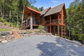 A two-story log cabin with a stone foundation, a stone retaining wall, and a gravel driveway. - Brand-New LUXE Creekside Cabin w Hot Tub, Fire Pit (Banner Elk)