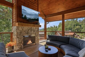 A wooden deck with a stone fireplace, a large TV screen, and a view of the surrounding forest. - Brand-New LUXE Creekside Cabin w Hot Tub, Fire Pit (Banner Elk)