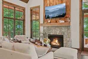 Ein gemütliches Wohnzimmer mit Kamin aus Stein, großen Fenstern und einem flachbildfernseher, der darüber angebracht ist.