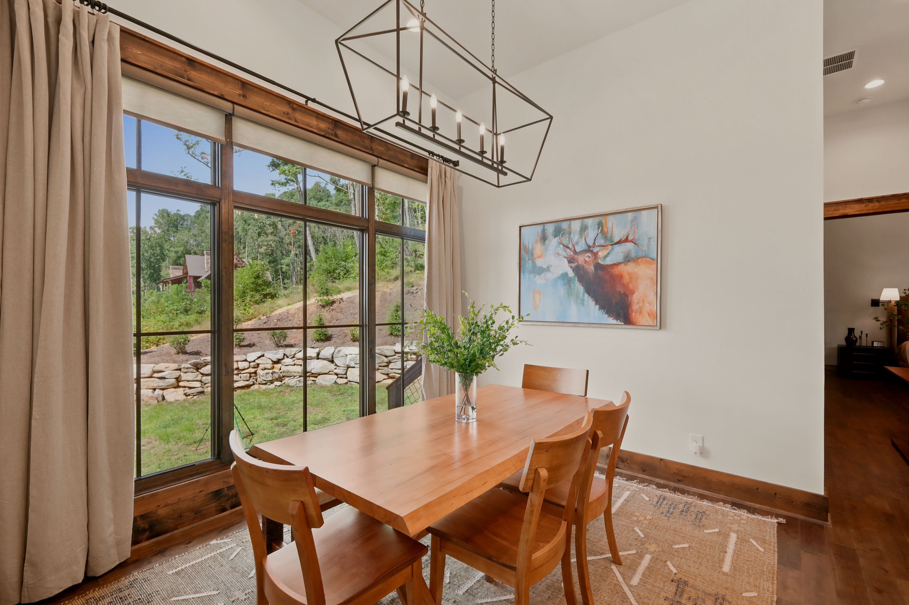 Un comedor con una mesa y sillas de madera, ventanas amplias con cortinas, una lámpara colgante y un cuadro en la pared.