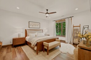 Quarto com cama de madeira, mesas de cabeceira, um banco, ventoinha no teto, janela com cortinas e um quadro na parede.