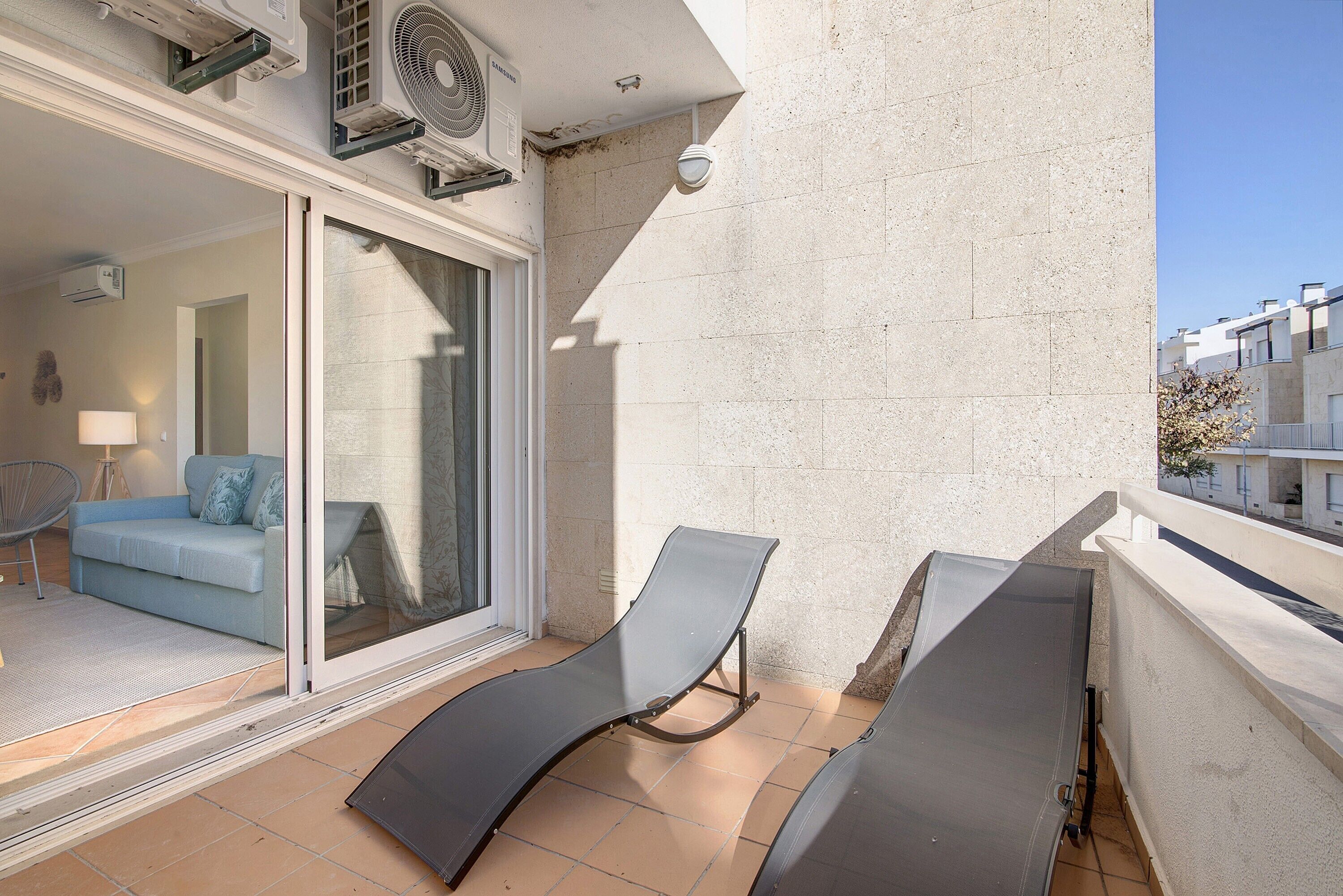 Ein Balkon mit zwei Lounge-Stühlen, eine Schiebetür zu einem Wohnbereich mit Sofa und Lampe sowie eine Wand-Klimaanlage.
