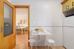 A kitchen with a white refrigerator, a small dining table with chairs, and a wooden door. - Apartment '1H Narciso' with Shared Pool, Wi-Fi and Air Conditioning (Santa Luzia)