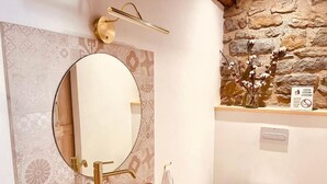 Une salle de bain avec un miroir rond, un lavabo blanc, un robinet doré et un mur en pierre.