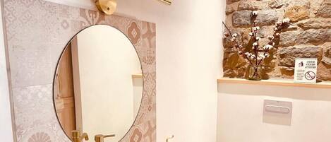 A bathroom with a round mirror, a white sink, a gold faucet, and a stone wall.