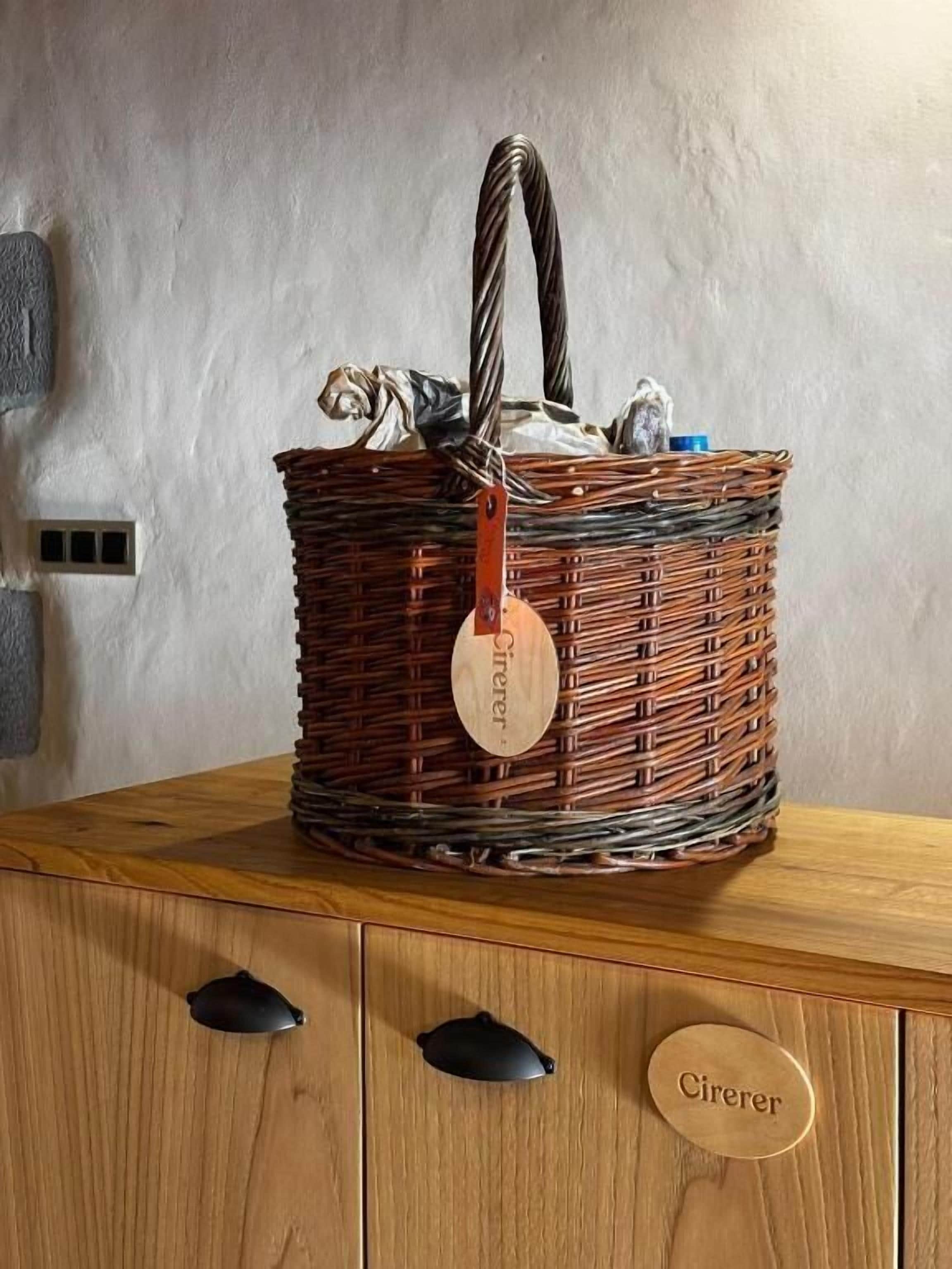 Una cesta de mimbre con asa metálica y una etiqueta de madera que dice 'Cirerer', sobre un mueble de madera con dos asas negras y una placa de identificación.