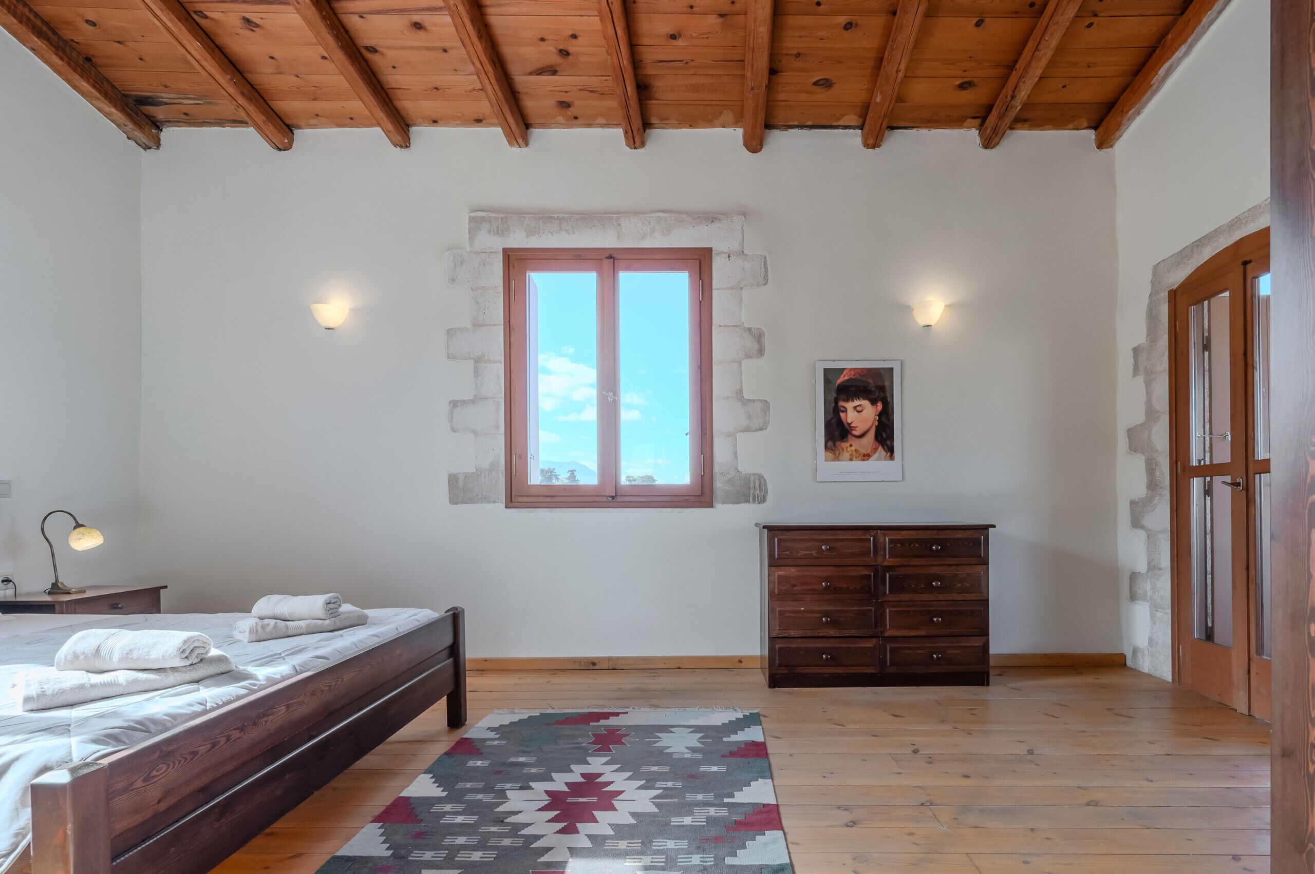 A bedroom with a wooden ceiling, a bed with neatly folded towels, a dresser, a window with a view, and a wall-mounted light.