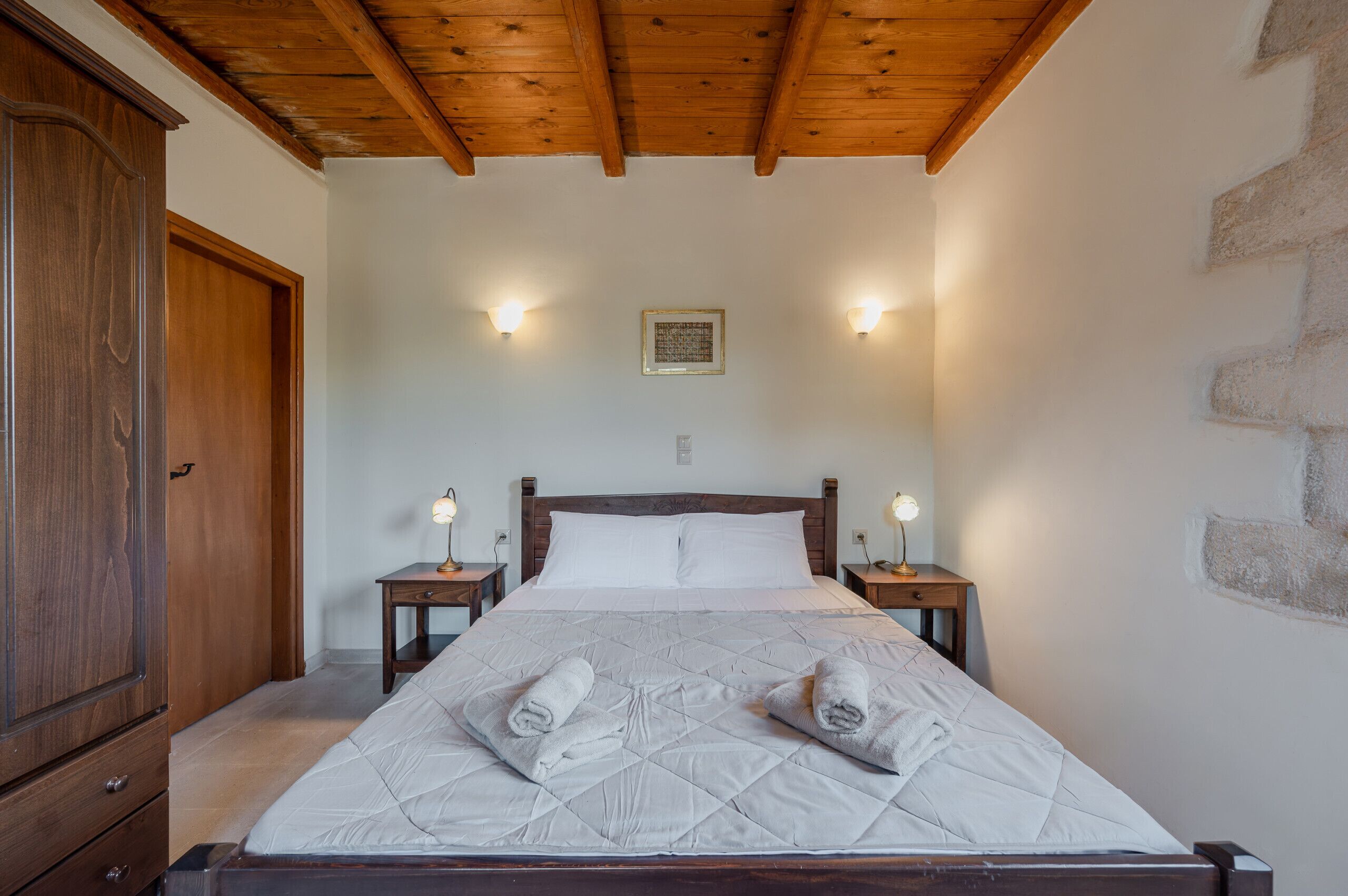A bedroom with a wooden ceiling, a large bed with white linens, two bedside tables with lamps, and a wooden wardrobe.