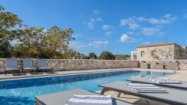 A swimming pool with lounge chairs and towels, a stone wall, and a building with a red roof in the background. - Vamos Villa (Apokoronas)