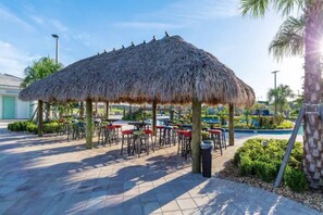 A thatched-roof outdoor seating area with multiple tables and chairs, surrounded by greenery and a pool in the background. - Modern Oasis at Storey Lake w/ 4BR & Heated Pool (Kissimmee)