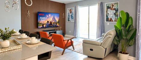 A modern living room with a flat-screen TV, a dining area with a table and chairs, and a large potted plant.