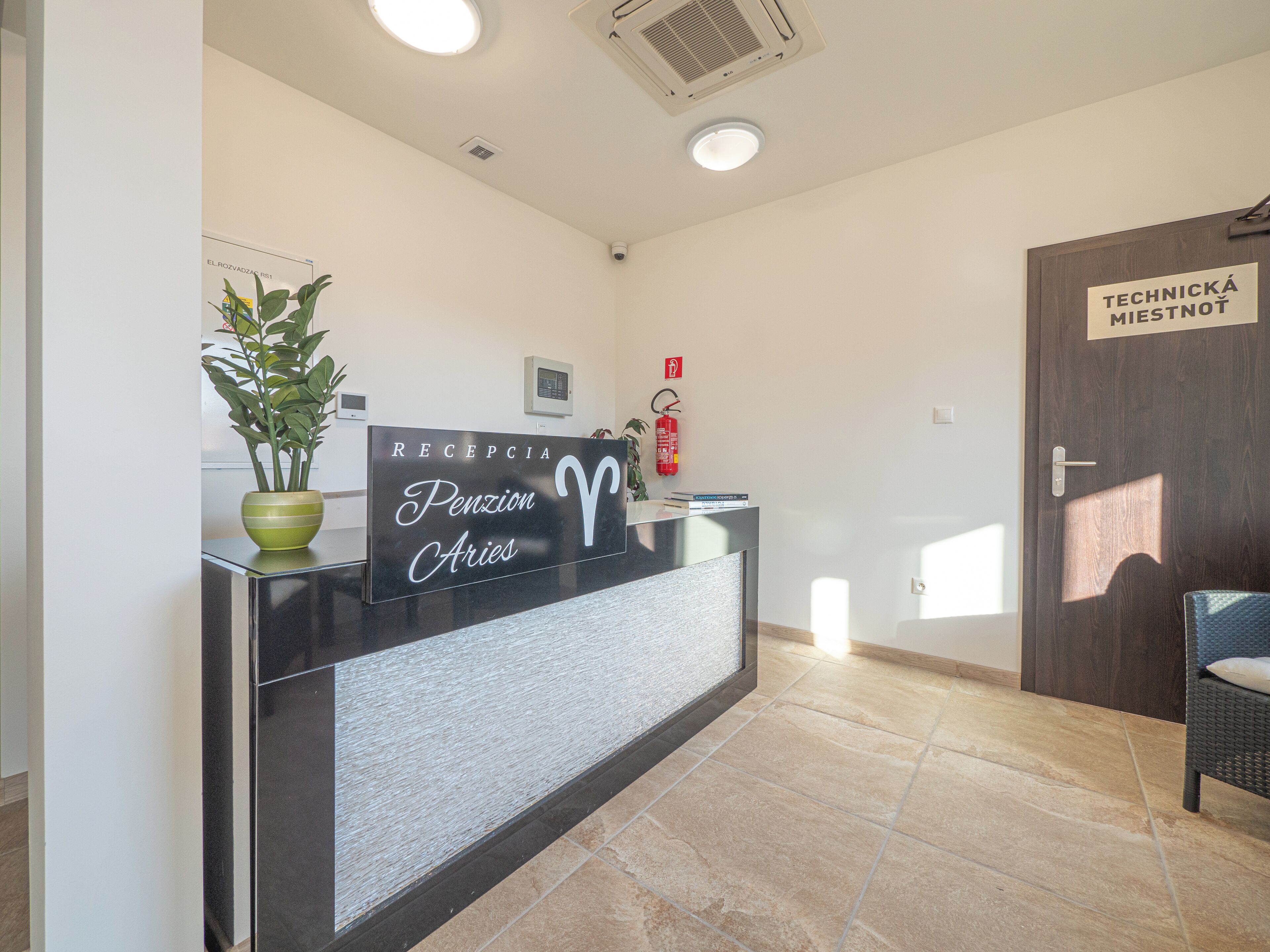 A reception desk with a sign reading "RECEPCIA Pension Aries," a potted plant, a fire extinguisher, and a door labeled "TECHNICKÁ MIESTNOT."