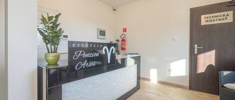A reception desk with a sign reading "RECEPCIA Pension Aries," a potted plant, a fire extinguisher, and a door labeled "TECHNICKÁ MIESTNOT."