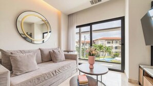 Een moderne woonkamer met een bank, een ronde spiegel, een klein rond tafeltje met een boek, en een groot raam met uitzicht op een gebouw en groen.