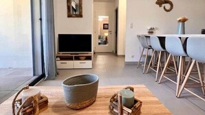 A modern living room with a large window, a flat-screen TV, a dining area with a table and chairs, and a decorative mirror on the wall. - Apartment in Maghjina near port and town center (Porto-Vecchio)