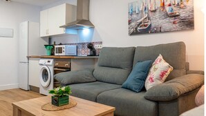 En moderne stue med en grå sofa, et træbord med en plante, en vaskemaskine, en mikrobølgeovn og et maleri af sejlbåde på væggen.