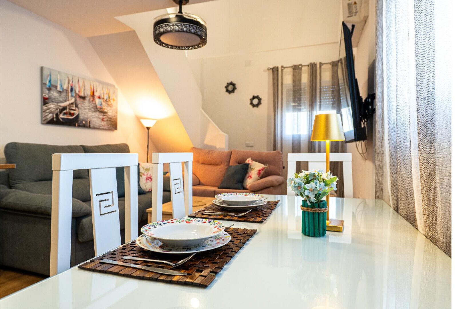 A dining area with a white table set for two, a green vase with flowers, and a painting of sailboats on the wall.