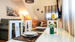 Et spiseområde med et hvidt bord dækket til to personer, en grøn vase med blomster og et maleri af sejlbåde på væggen.