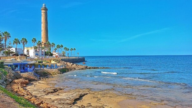 Un paysage côtier avec un phare imposant, une côte rocheuse, des palmiers et un bâtiment doté d’espaces de repos bleus.
