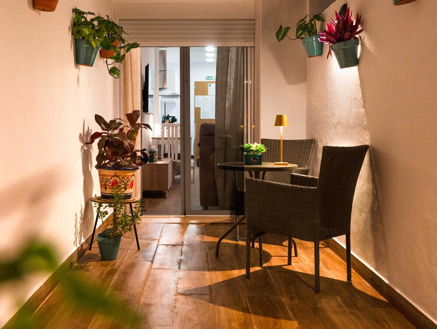 A narrow hallway with a wooden floor, a wicker chair, a small table with a lamp, and potted plants.