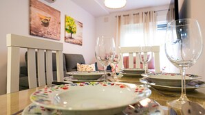 Een eettafel gedekt met borden, glazen en bestek, een witte stoel en een bank in een kamer met een plafondlamp en twee schilderijen aan de muur.