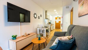 Een moderne woonkamer met een grijze bank, een klein rond tafeltje, een aan de muur bevestigde televisie en een keuken op de achtergrond.