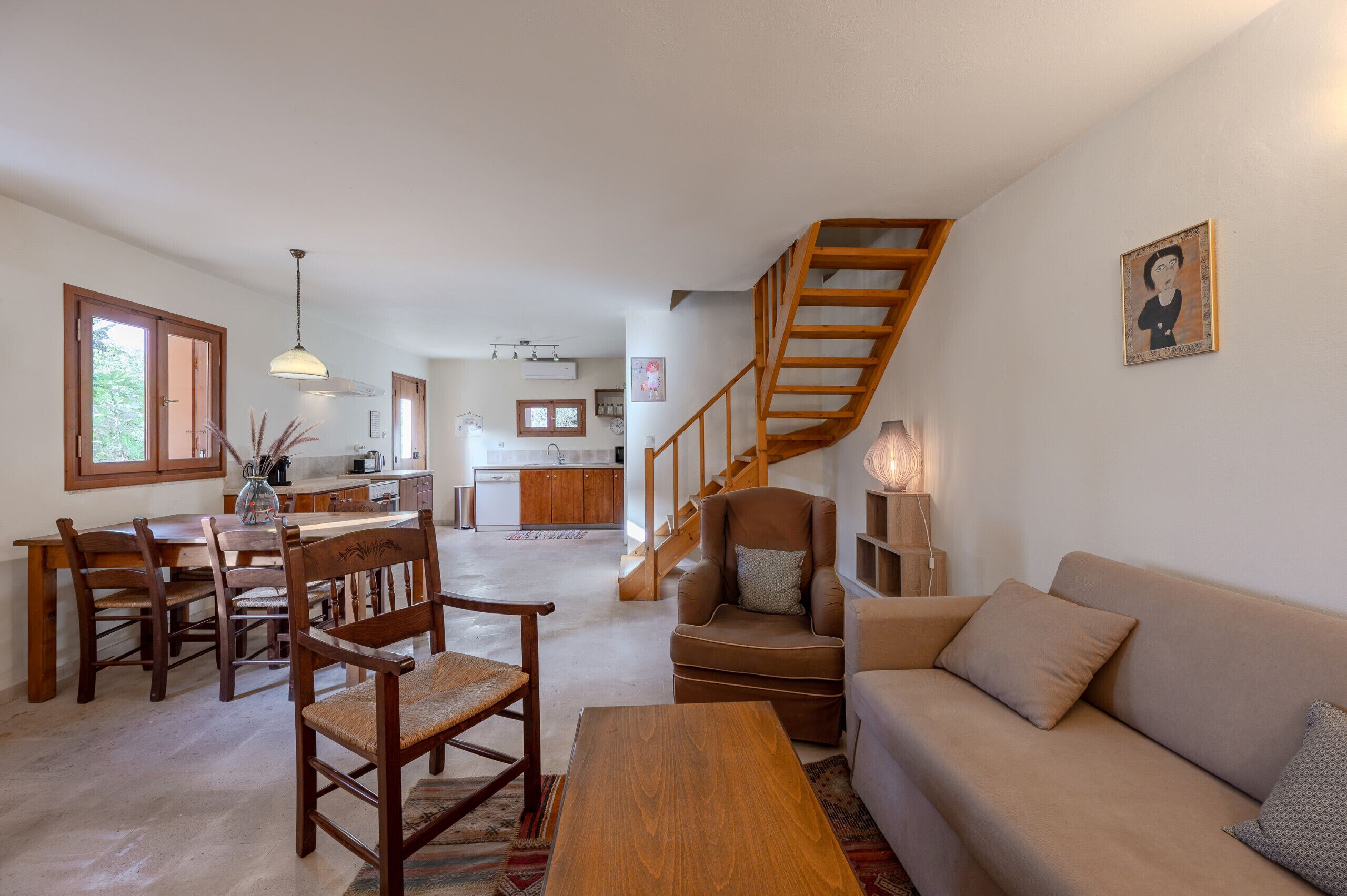 A spacious living area with a wooden staircase, a dining table with chairs, a sofa, and a kitchen in the background.