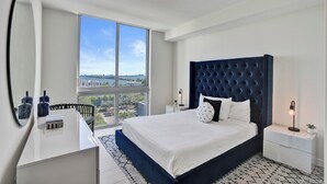 Un dormitorio moderno con una cama grande, una mesita de noche, un espejo y vistas a la ciudad a través de amplias ventanas.