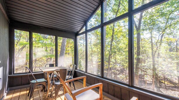 Un espacio de estar techado al aire libre con una mesa y sillas, ventanas amplias y vistas a un bosque.