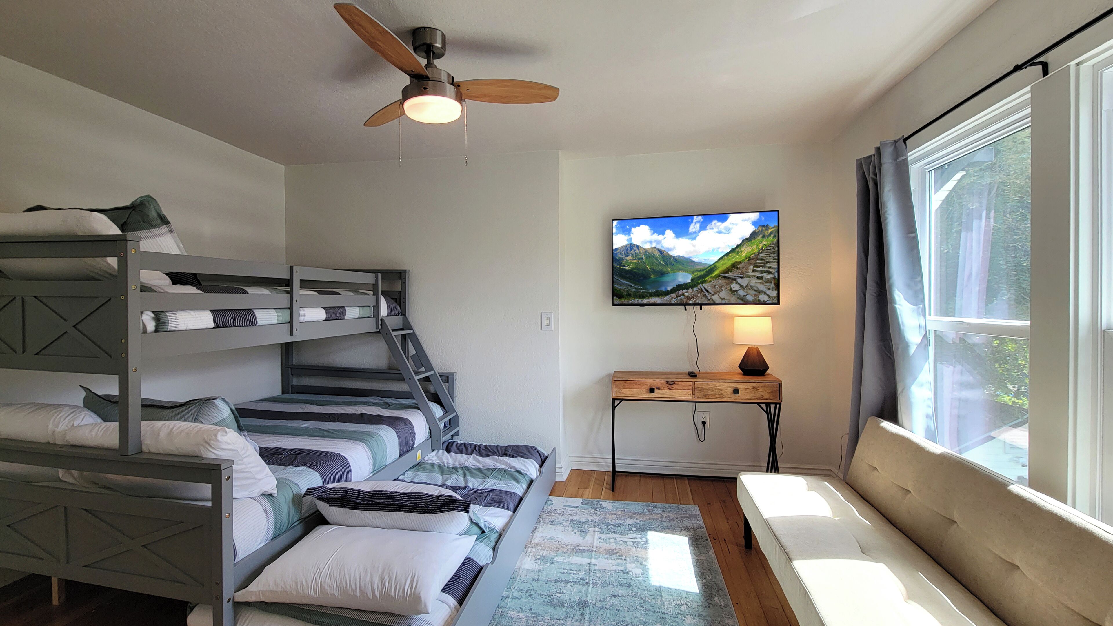 A room with bunk beds, a desk with a lamp, a television on the wall, and a window with curtains.