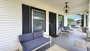 Eine überdachte Veranda mit einem grauen Sofa, zwei Stühlen, einer schwarzen Tür und einem weißen Geländer.