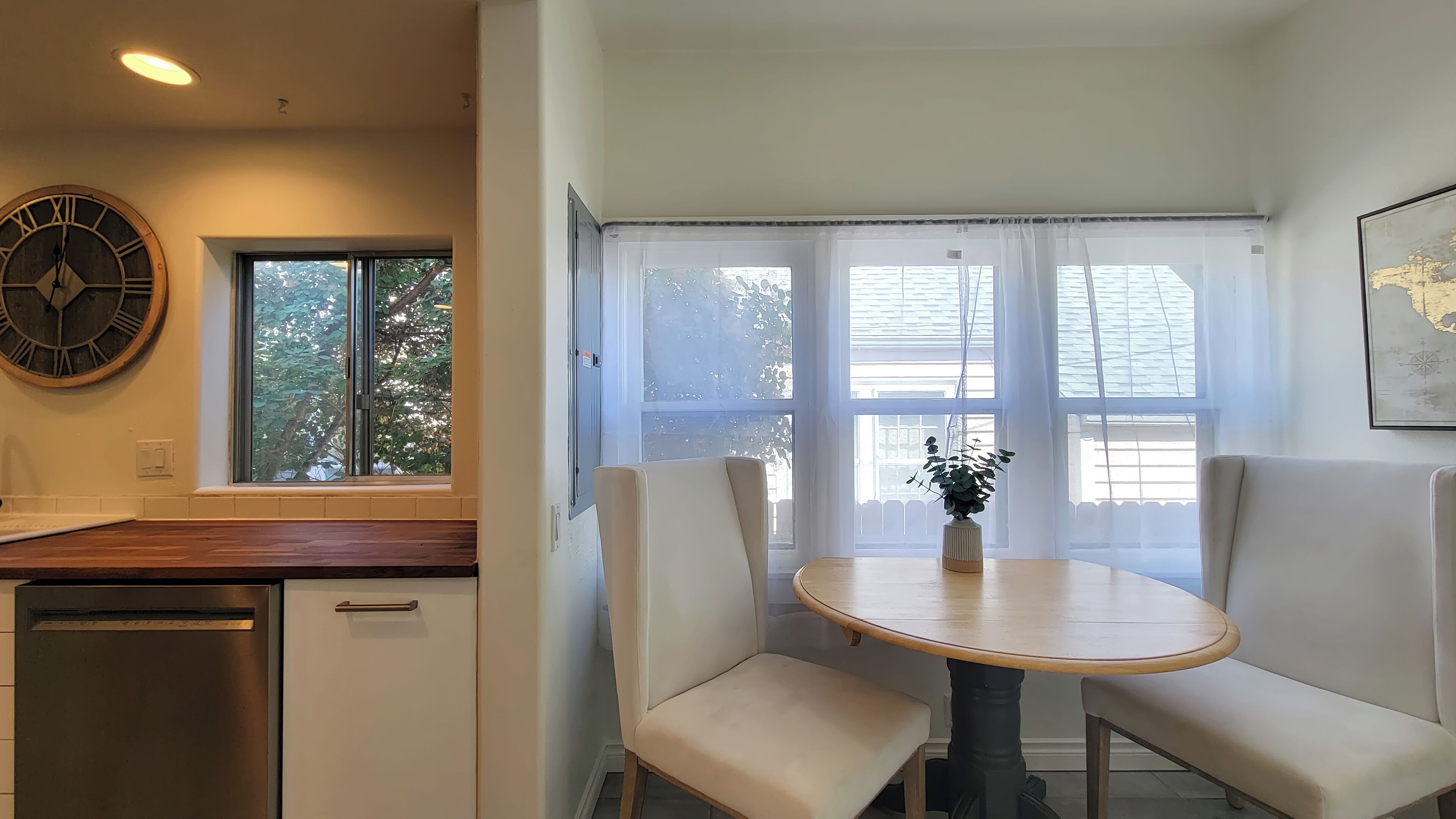 Una cocina compacta con lavavajillas integrado, una mesa redonda de madera con dos bancos acolchados y un ventanal con cortinas de encaje.