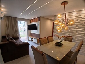 A modern living room with a flat-screen TV, a dining table with chairs, a sofa, and a decorative light fixture. - Apts in a Condomin. w/ Complete Leisure Facilities, 450 m fr/ the Beach - Vars (Ilhéus)