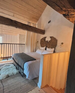 Um quarto aconchegante com teto de madeira, cama com lençóis brancos, tapete fofo e cabeceira rústica.