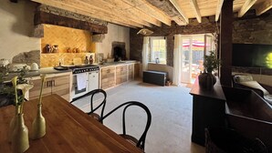 A rustic kitchen with a stone wall, wooden beams, and a built-in oven. There is a dining table with chairs and a TV mounted on the wall. - Domaine du Tertre ychot (Dol-de-Bretagne)