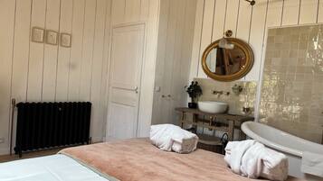 A bathroom with a white door, a wooden vanity with a sink, a mirror, and towels on the bed.