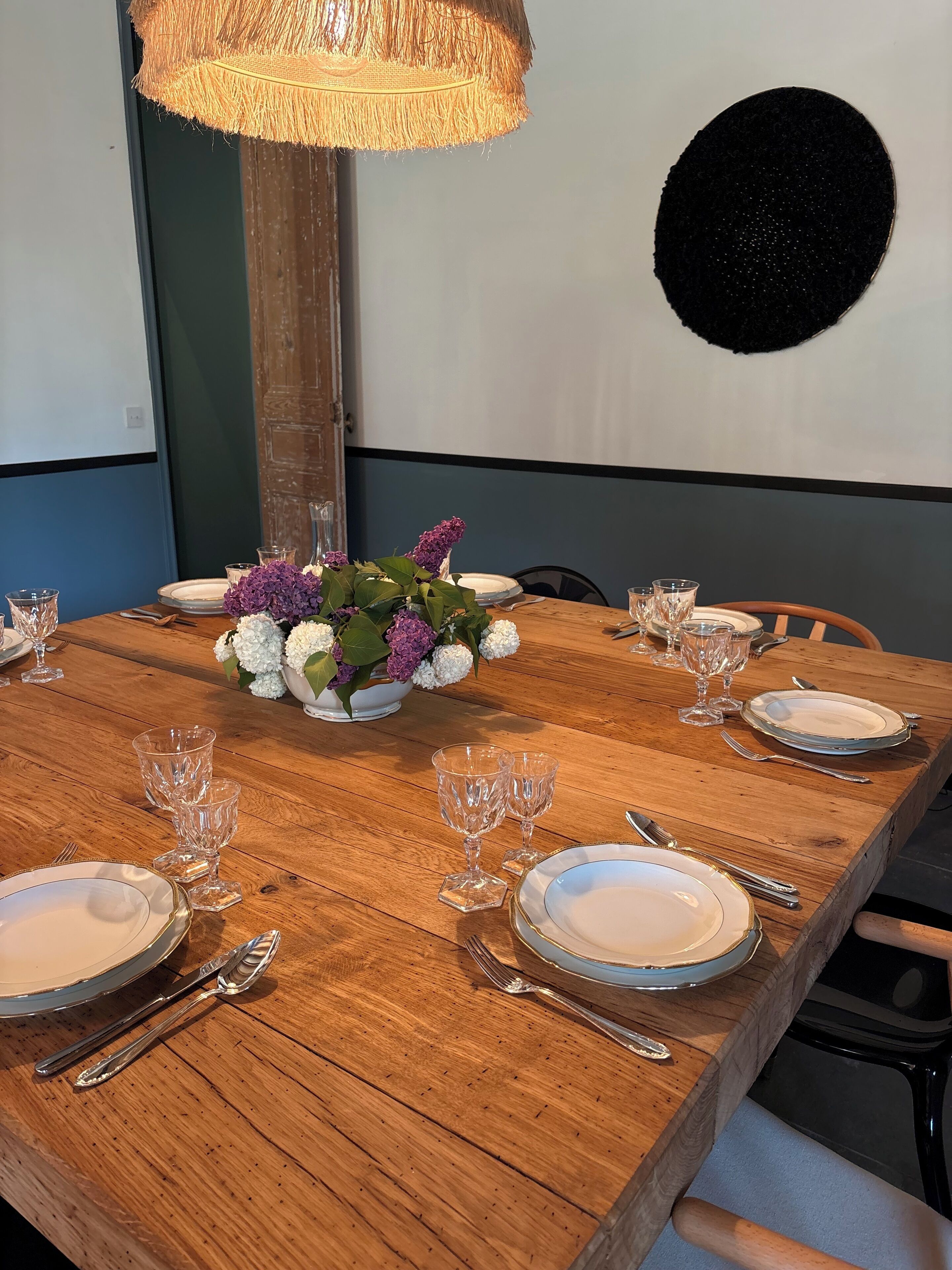 Une table à manger en bois est dressée avec de l’argenterie, de la vaisselle blanche et de la verrerie transparente. Un luminaire tressé est suspendu au-dessus, et un arrangement floral orne le centre de la table.