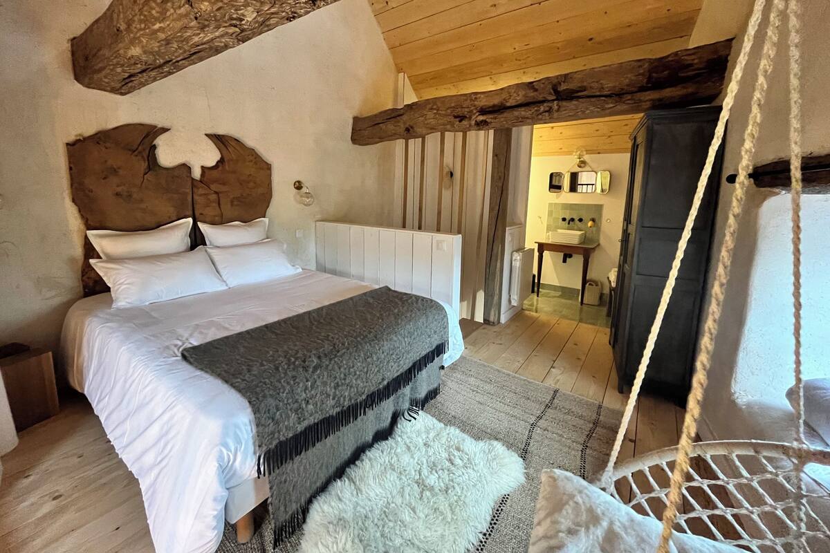 A bedroom with a large bed, wooden beams, a hanging chair, and a view of a kitchen area.