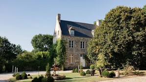 Un bâtiment en pierre doté d’un toit en pente, un jardin bien entretenu avec divers arbustes et une fontaine centrale, ainsi qu’une allée de gravier menant à l’entrée.