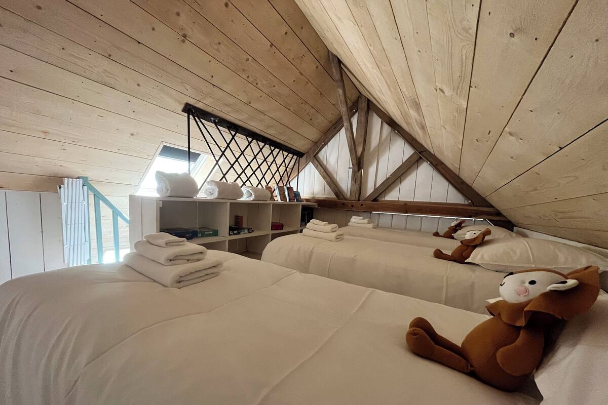 A cozy attic bedroom with two beds, a wooden ceiling, and a shelf with towels and toiletries.