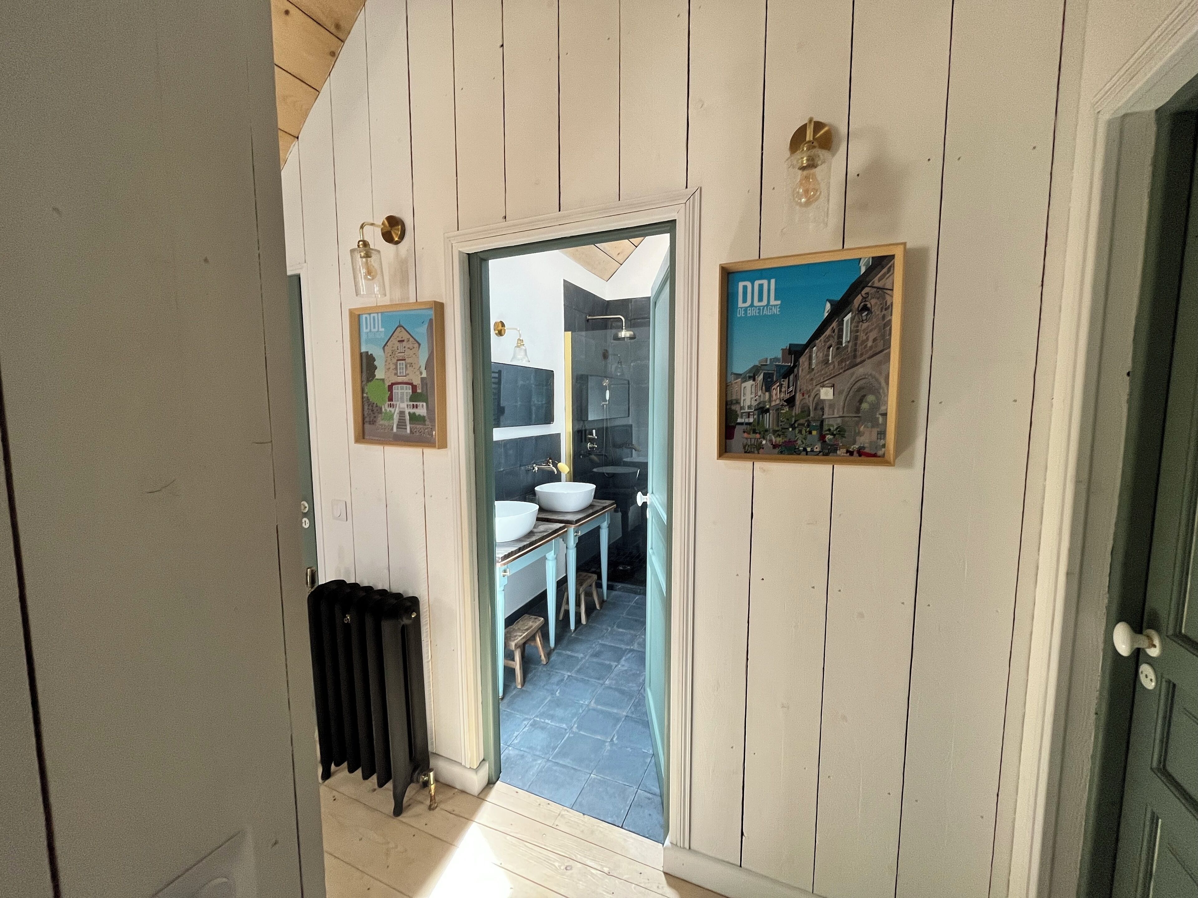 Een smalle gang met houten lambrisering, een radiator, een lijst met een afbeelding van een gebouw en een lijst met een straatbeeld.