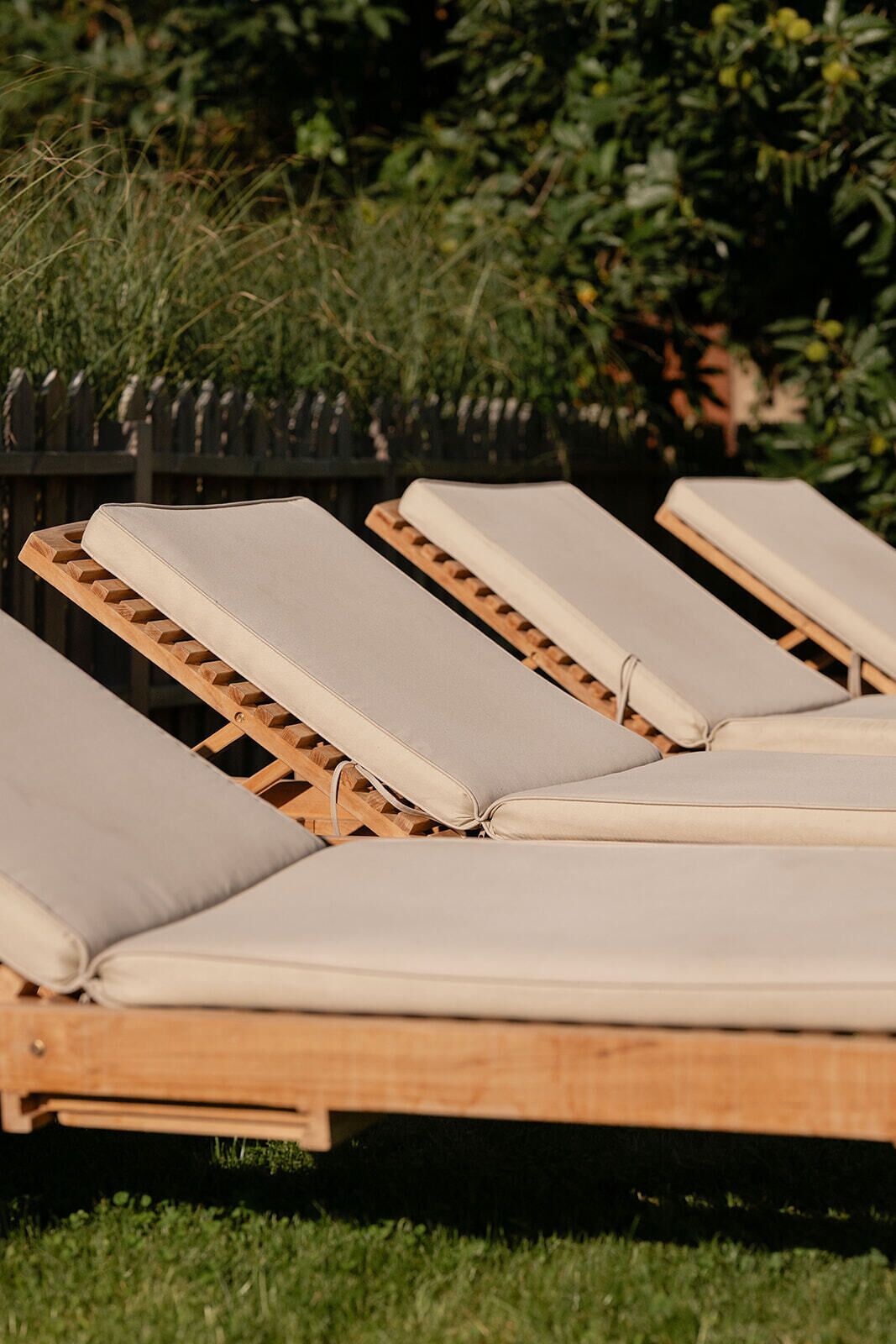 Une rangée de fauteuils de jardin en bois, dotés de coussins beiges, est disposée à l’extérieur, avec une clôture en bois et de la verdure en arrière-plan.