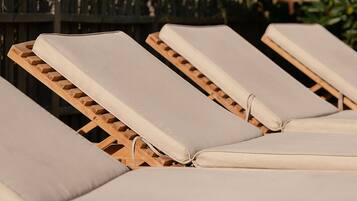 Une rangée de fauteuils de jardin en bois, dotés de coussins beiges, est disposée à l’extérieur, avec une clôture en bois et de la verdure en arrière-plan.