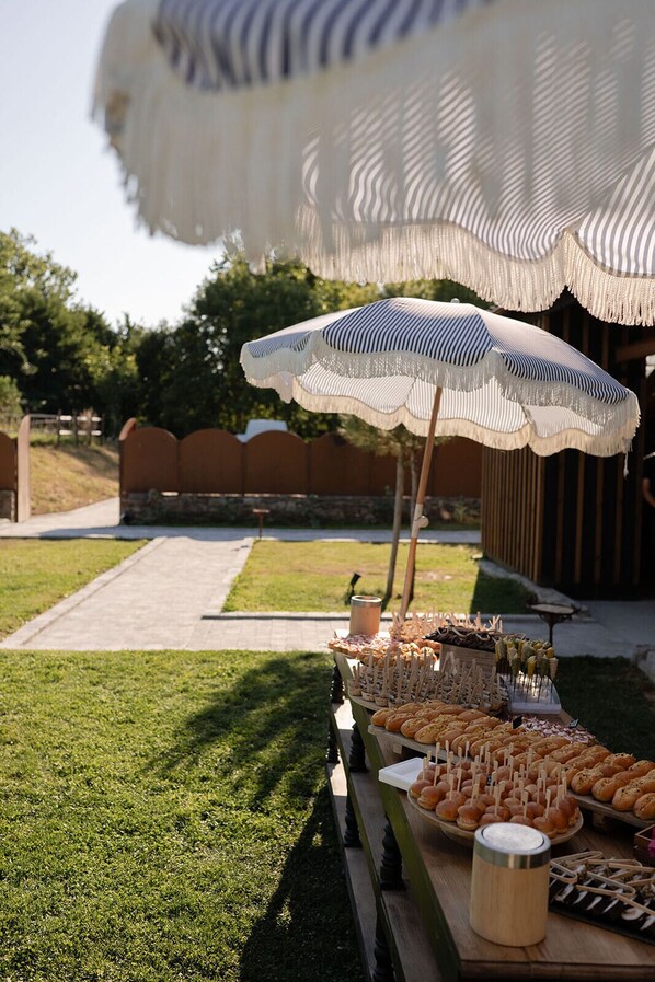 An outdoor buffet setup with a variety of food items, including skewers and pastries, under a large striped umbrella. - Domaine du Tertre ychot (Dol-de-Bretagne)