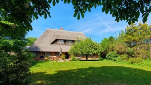 Une maison traditionnelle avec un toit de chaume, entourée d’une pelouse bien entretenue et d’arbres.