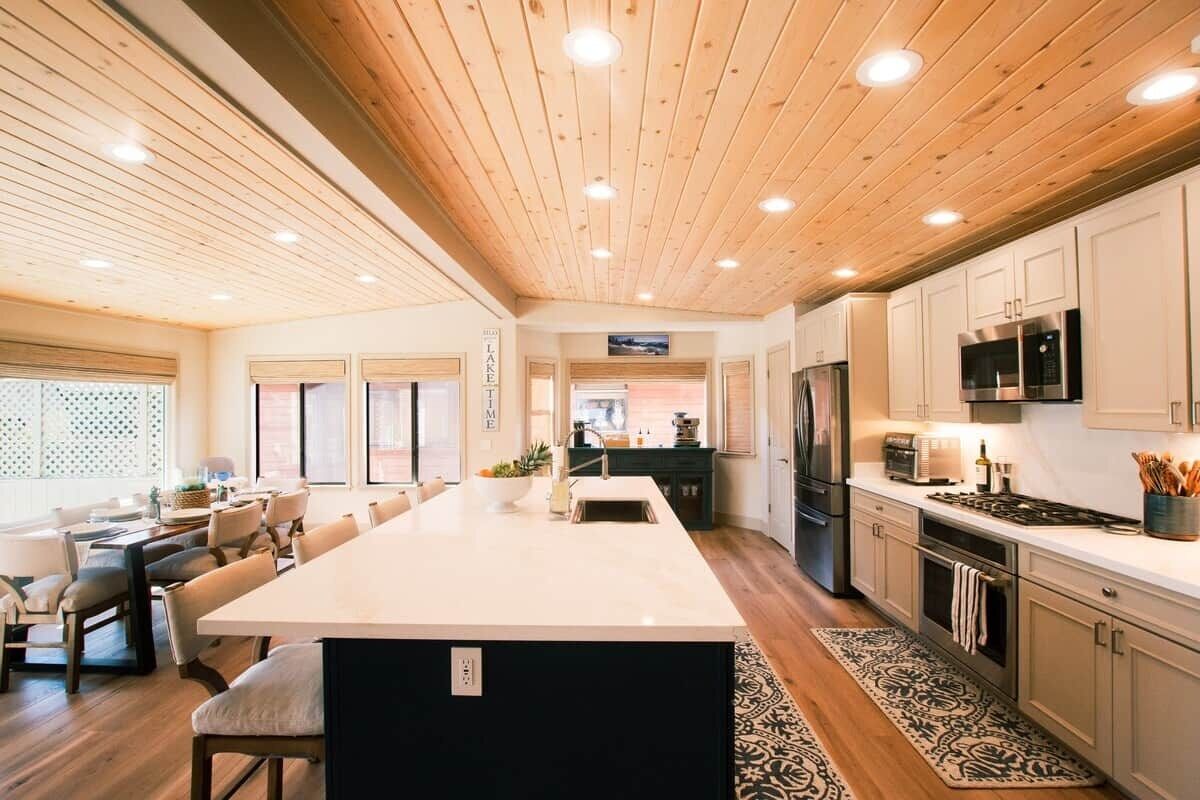 A modern kitchen with wooden ceiling, white cabinets, a central island, and a dining area with chairs.